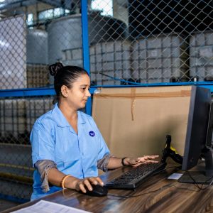 A woman in blue uniform operates a computer inside a factory warehouse, focusing on her task.